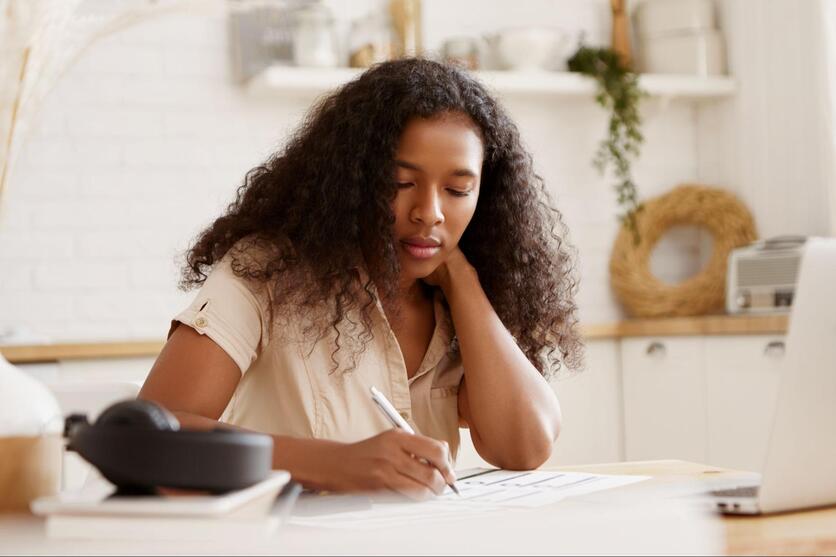 Mulher negra, sentada em frente a um notebook, fazendo anotações com uma expressão séria.