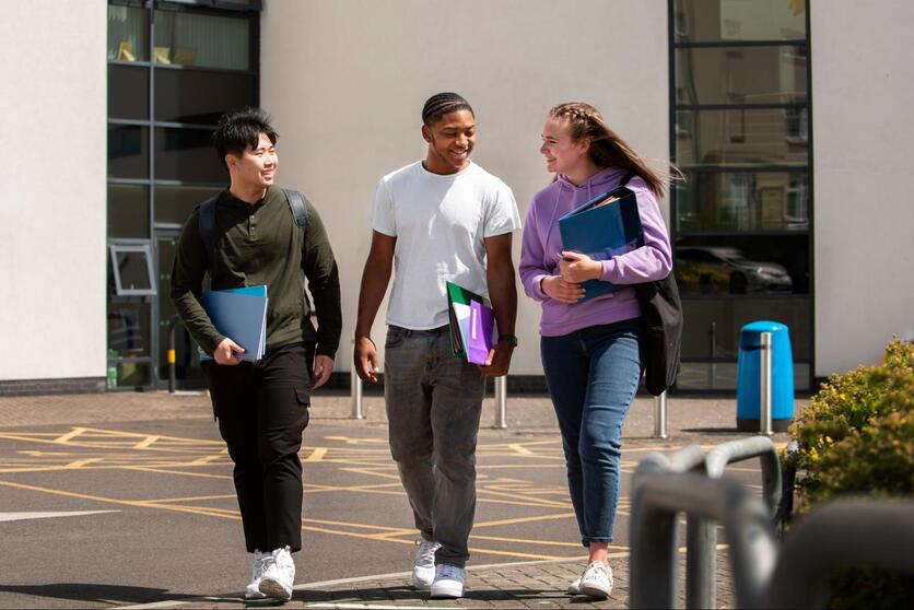 Três jovens, lado a lado, caminhando na calçada com materiais de estudos nas mãos.