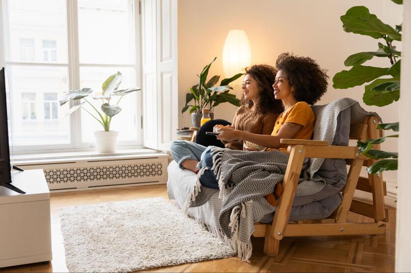Duas mulheres sorridentes e descontraídas sentadas no sofá da sala de estar, desfrutando de um momento de relaxamento em frente à televisão.