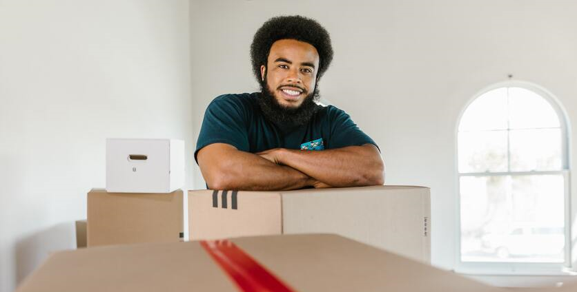 Homem negro, sorrindo, se apoiando em cima de uma caixa de papelão. Ao seu redor, há diversas outras caixas, indicando que está se mudando.