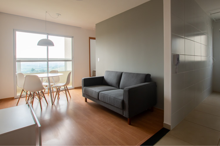 Sala de estar de apartamento mobiliado Luggo, com mesa, sofá e rack. À esquerda, é possível ver uma janela com sacada.
