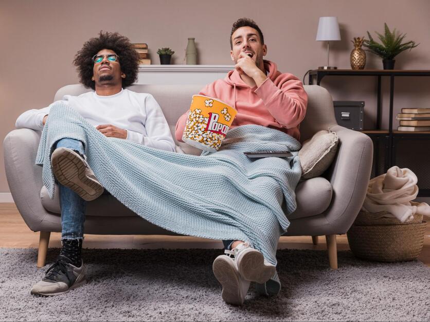 Dois jovens sentados num sofá bege, com um cobertor e um balde de pipoca no colo.