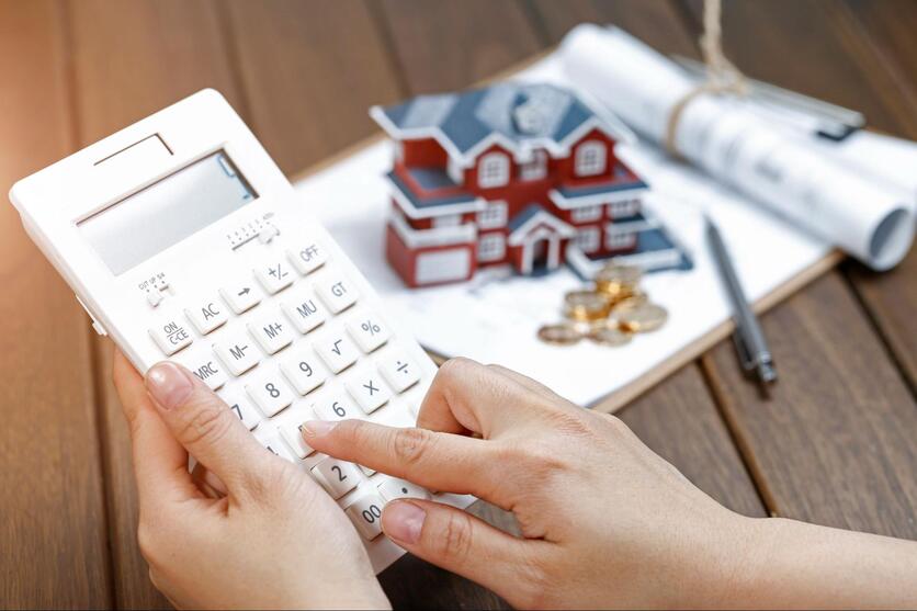 Mão feminina digitando em uma calculadora, com uma prancheta, miniatura de uma casa e moedas ao fundo.