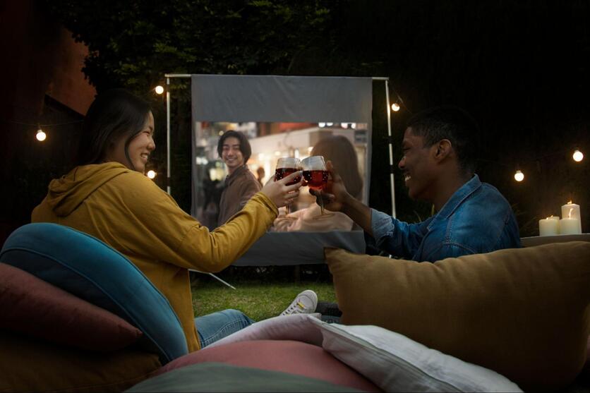 Casal de jovens brindando taças de champanhe, sentados em um gramado com um projetor de filme à sua frente.