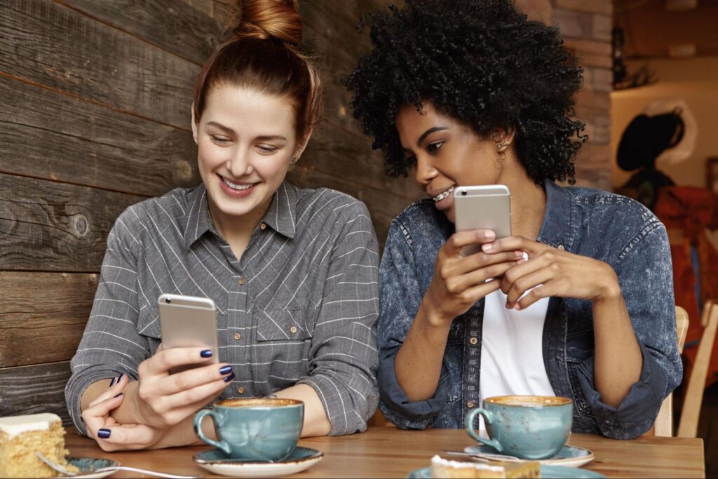 Casal de mulheres em frente a uma mesa de café, sorrindo enquanto olham para seus celulares.