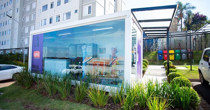 Área externa do Luggo Cenarium, com loja de conveniência 24hrs cercada por grama verde. Ao fundo, um conjunto de cestas de coleta seletiva de lixo.