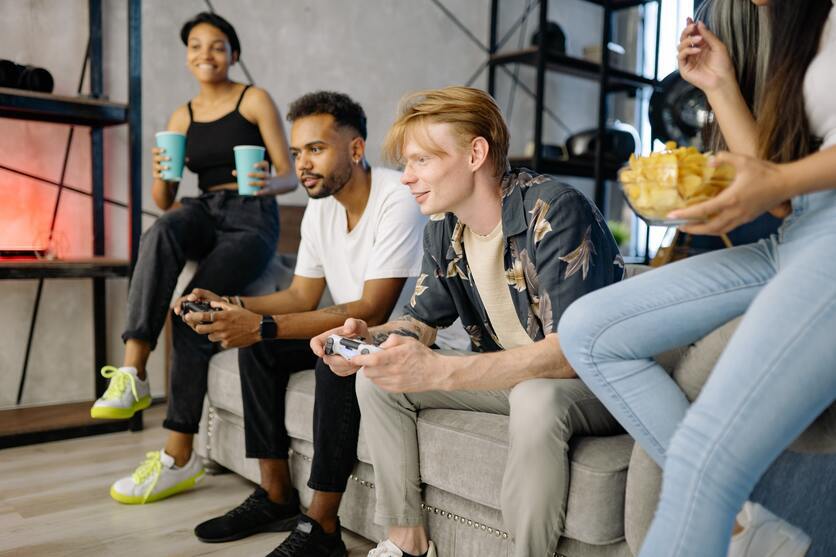 Grupo de jovens sentados em um sofá, com controles de videogame, copos e salgadinhos nas mãos.
