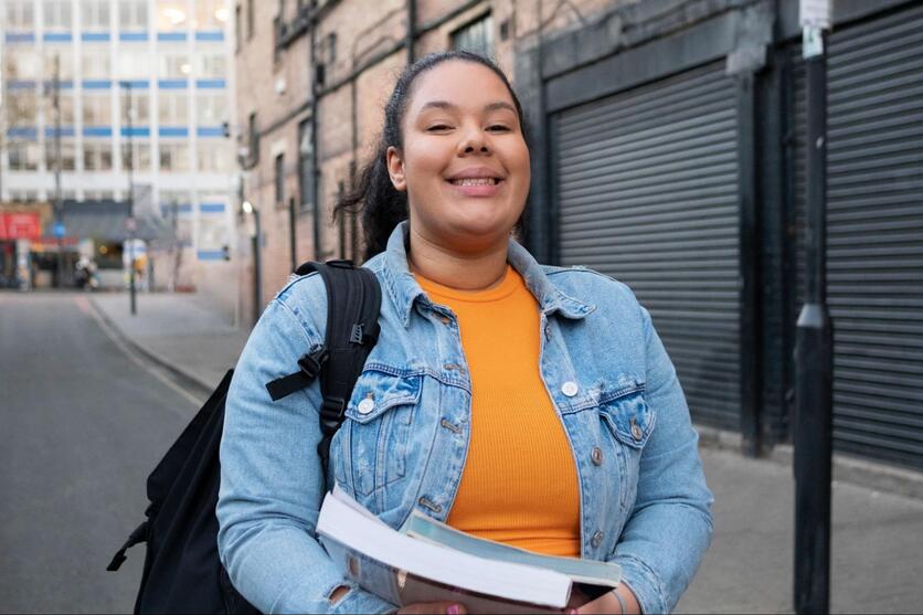 Mulher negra sorrindo enquanto caminha numa rua, segurando dois livros na mão e com uma mochila nas costas.