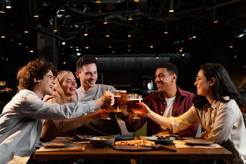 Grupo de amigos sentados em uma mesa de restaurante, sorrindo, enquanto fazem um brinde com copos de cerveja/chopp.