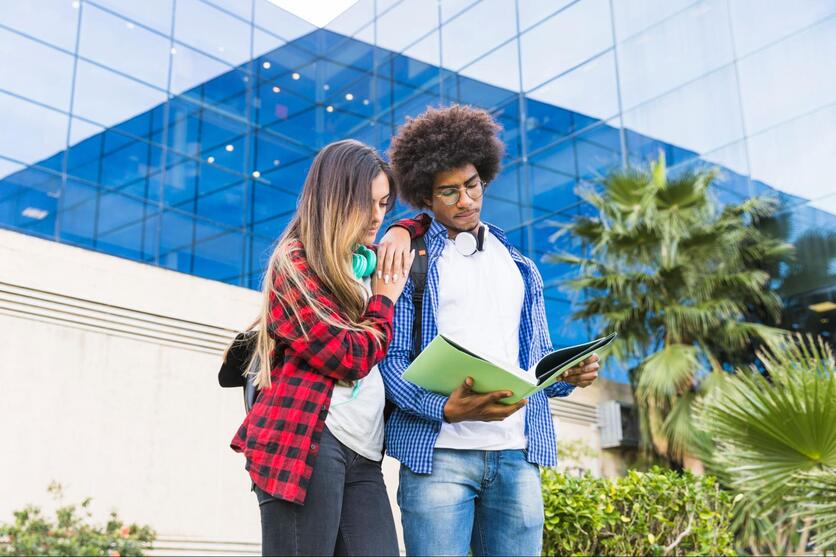 Dois jovens, uma mulher e um homem, em frente a um prédio com um livro aberto e fones de ouvido no pescoço.