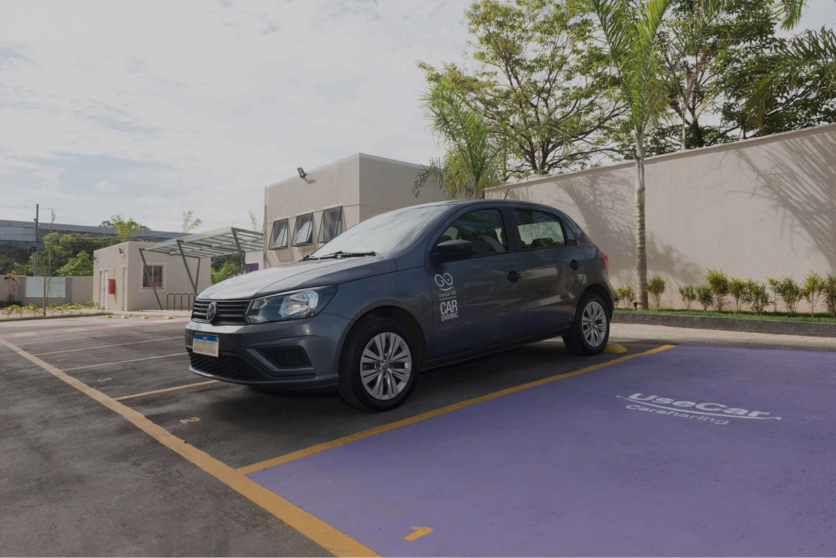 Carro cinza, em estacionamento Luggo e o logo da UseCar GO na porta. À direita, uma vaga pintada de roxo também com o logo da UseCar.