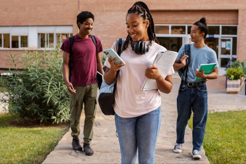 Três estudantes, caminhando em frente a um campus, sorrindo com mochilas nas costas e carregando livros nas mãos.