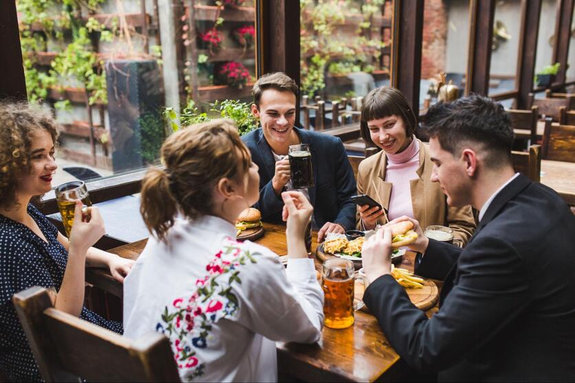 Grupo de amigos sentados em volta de uma mesa de restaurante, rindo e conversando, durante o dia.