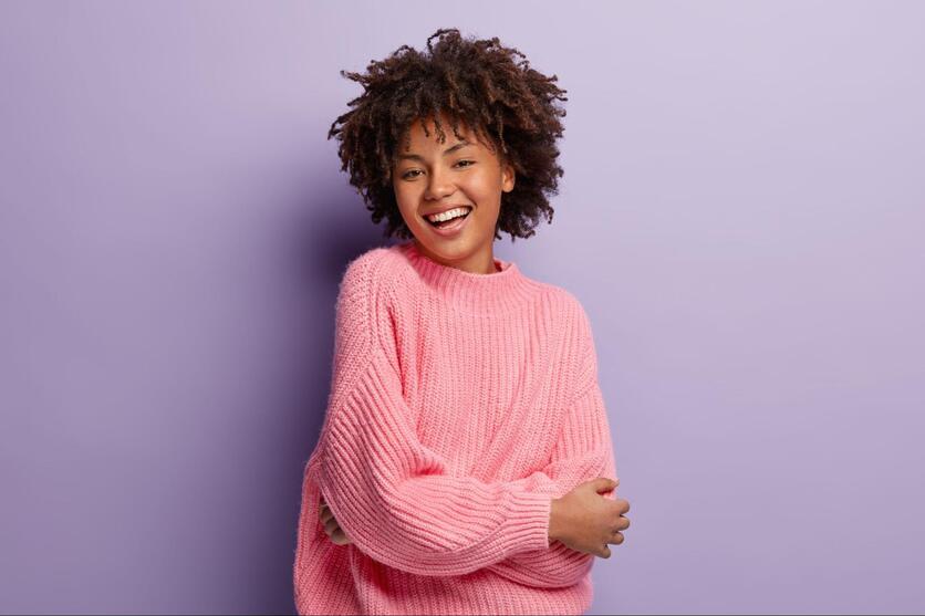 Jovem negra sorrindo, olhando para a câmera, vestindo um suéter rosa num fundo roxo, remetendo à Luggo.