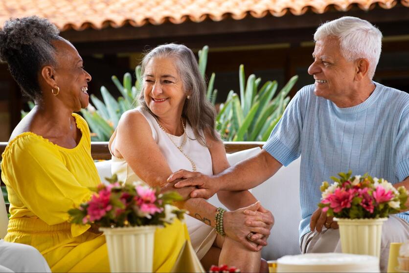 Três idosos conversando, sentados em sofá branco. Flores rosas em vaso a frente e, ao fundo, telhado e plantas.