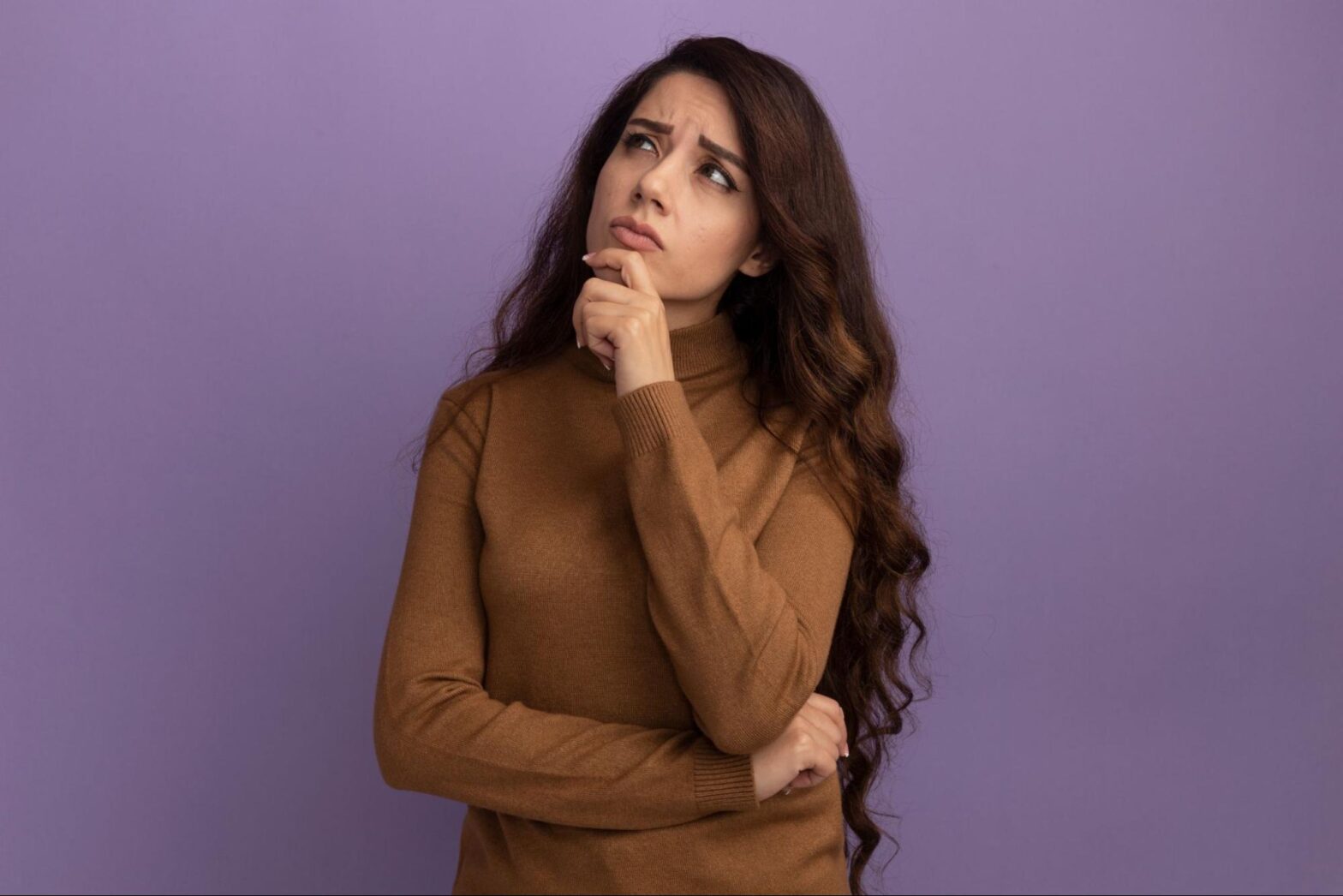 Mulher adulta, com blusa marrom, com a mão apoiada no queixo e olhando para cima com uma expressão pensativa.