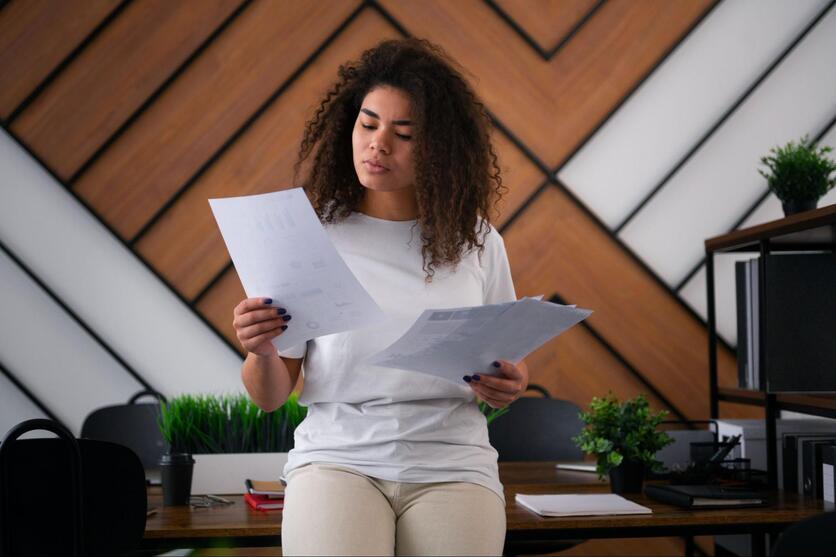 Mulher negra com uma expressão pensativa, em cima de uma mesa de escritório, com diversos documentos nas mãos.