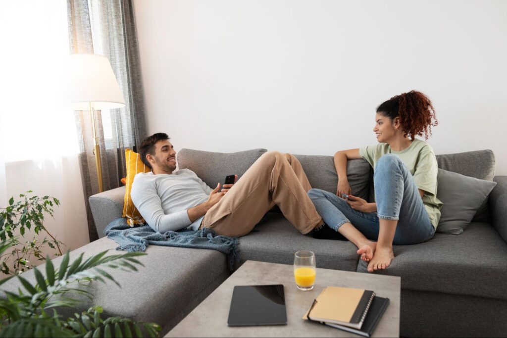 Casal sentado em sofá de apartamento, sorrindo enquanto conversam, relaxados.