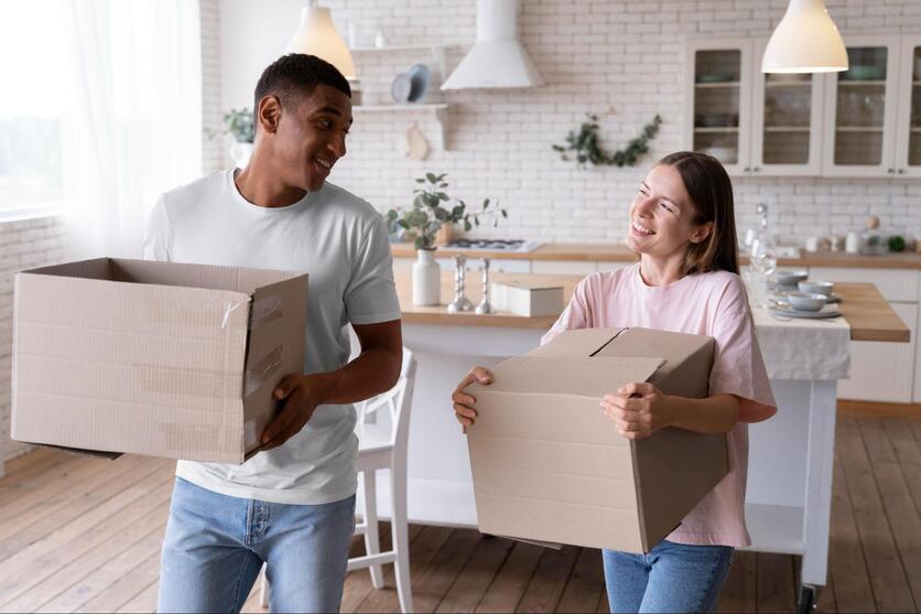 Um homem e uma mulher seguram caixas de mudança e se olham sorrindo, ao fundo é possível ver uma cozinha mobiliada e organizada.