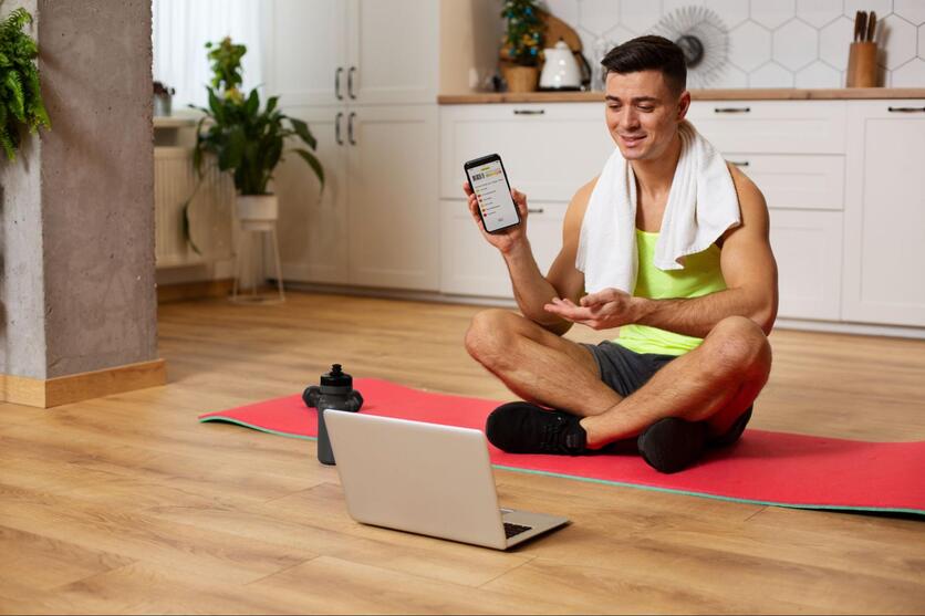 Homem sentado em um tapete de exercícios vermelho, sorrindo com um celularna mão. Na frente dele, há um notebook e uma garrafa de água.