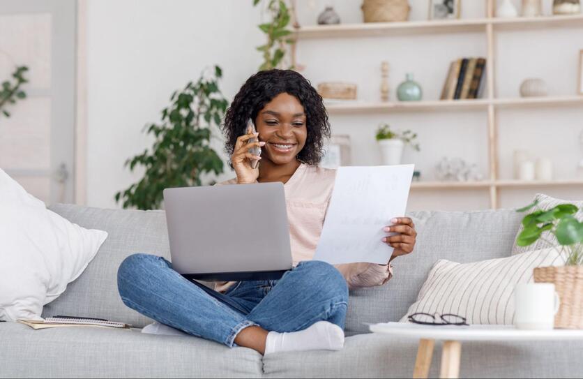 Mulher sorrindo, sentada no sofá, com o notebook sobre as pernas cruzadas, olhando para uma folha de papel e falando ao celular. Ao fundo, uma estante de madeira com itens de decoração e plantas.