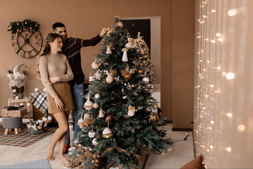 Um homem e uma mulher estão abraçados em frente a uma árvore de natal, o homem tem um enfeite na mão e eles aparentam estar decorando a árvore. A parede está decorada com luzes e atrás deles tem uma pequena mesa e bancos com ornamentos natalinos.