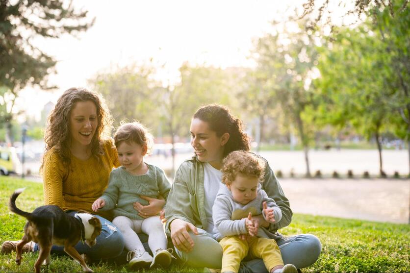 Duas mulheres sentadas na grama, lado a lado, cada uma tem uma criança pequena sentada em seu colo, um cachorro brinca ao lado deles. O ambiente é um parque ao ar livre.