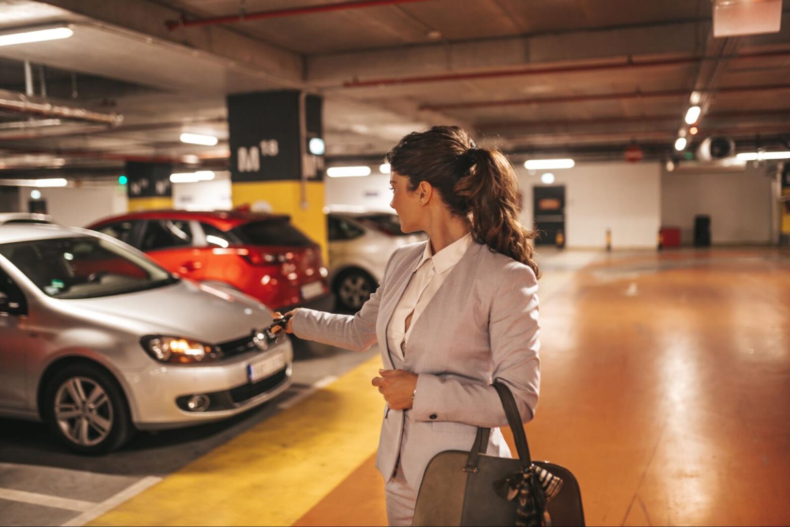 Mulher em uma garagem em condomínio, ela está com o corpo voltado para trás, acionando o alarme de um carro que está estacionado