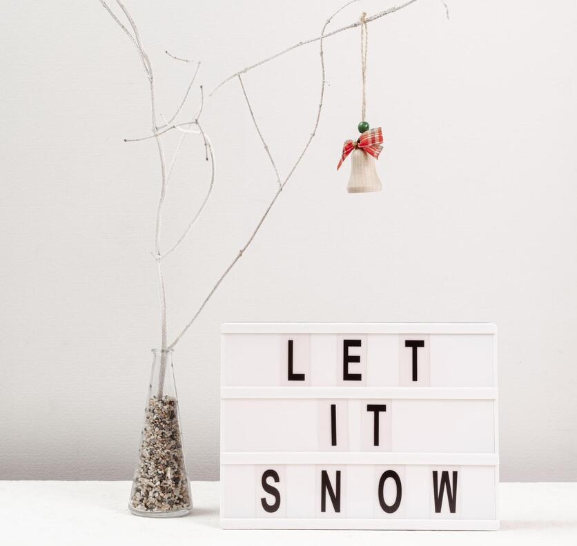 Decoração de natal minimalista. Placa de mesa branca, com letras pretas, com a frase “Let it snow”. Vaso de vidro transparente e em formato de funil, cheio de pequenas pedras, dele saem galhos sem folhas, em um dos galhos está pendurado um pequeno ornamento de natal.