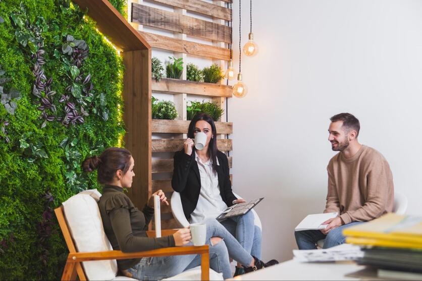 duas mulheres e um homem estão sentados e aparentam estar conversando. Eles têm canecas de louça e papéis nas mãos. O ambiente é decorado com plantas e é possível ver uma mesa com papéis.