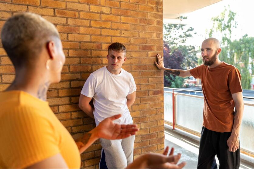 Uma mulher e dois homens conversam em um corredor. A mulher está de costas e com as mãos levantadas, como se estivesse argumentando. Os dois homens estão encostados em uma parede e olham a mulher com atenção. Ao fundo é possível um parapeito de vidro e folhagens de árvores.