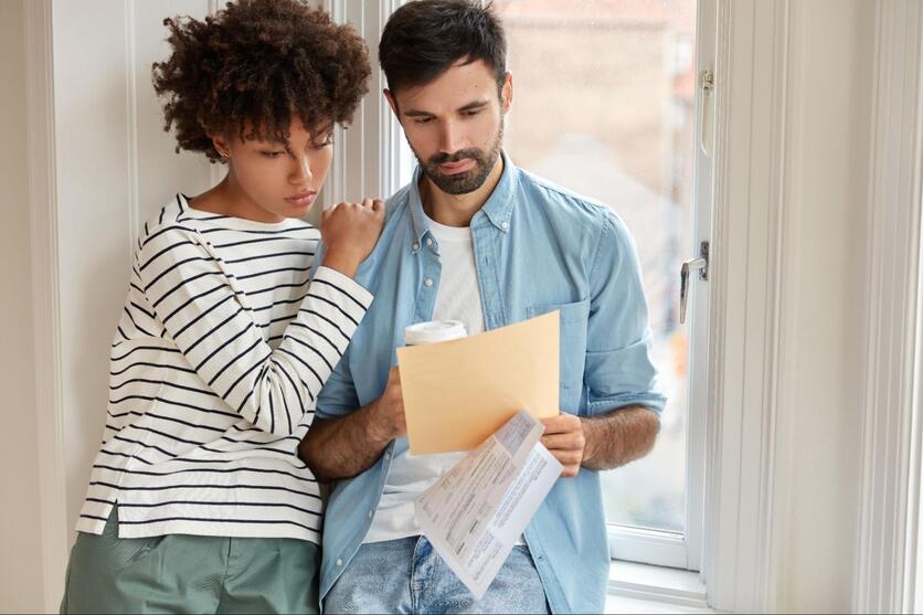 Um homem e uma mulher estão analisando papéis. A mulher está apoiada no ombro do homem e os dois estão encostados em uma janela de vidro. O homem segura um copo de café. Os dois tem expressões concentradas.