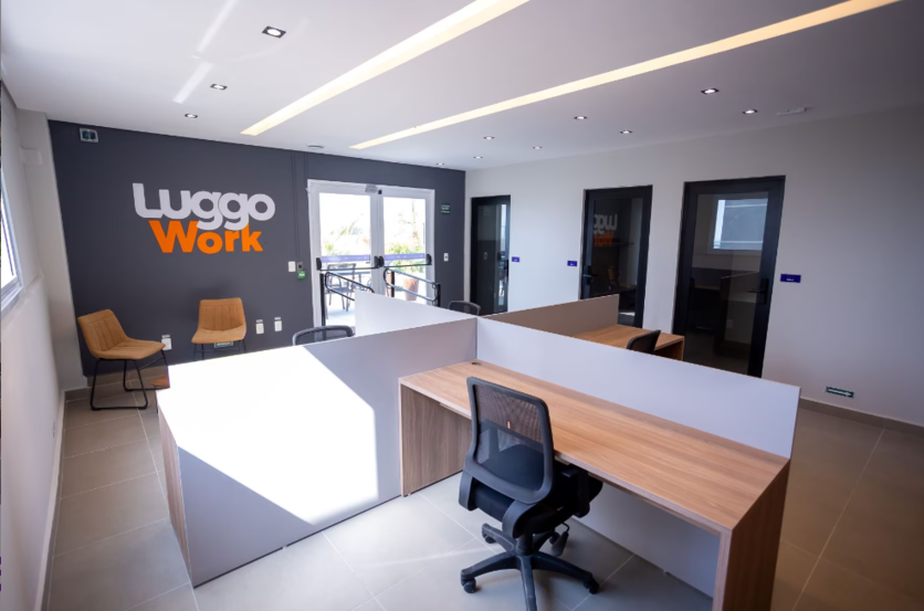 Espaço de coworking no condomínio da Luggo. O ambiente apresenta mesas de trabalho em madeira clara, cadeiras pretas e uma parede com o logo "Luggo Work" em destaque. Grandes janelas permitem a entrada de luz natural.